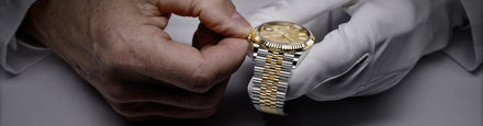 A gloved hand holds a luxury gold and silver wristwatch as another hand adjusts its bezel.