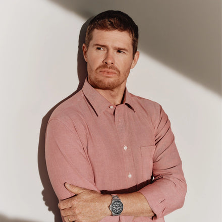 Man in a pink button-up shirt with arms crossed, standing against a wall with dramatic shadow.