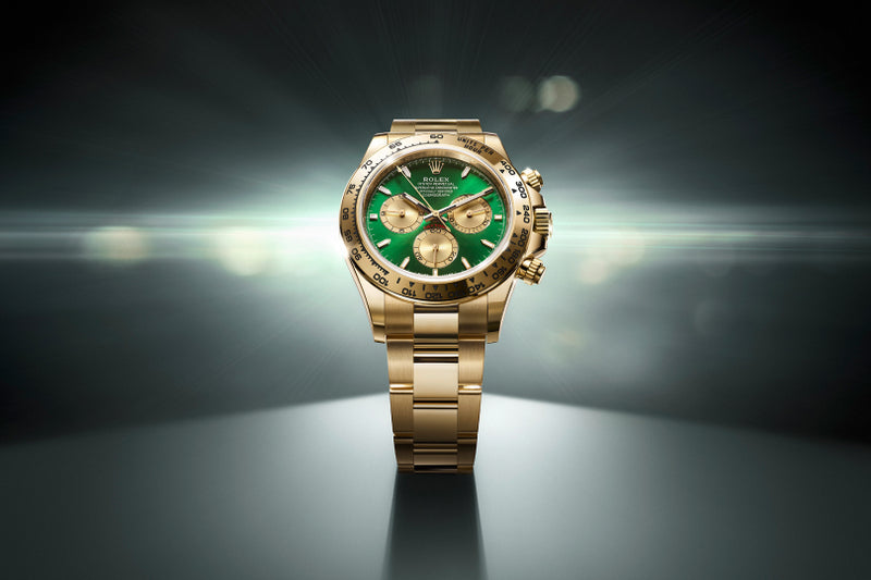 Gold wristwatch with a green face and three subdials, illuminated on a dark, spotlighted background.