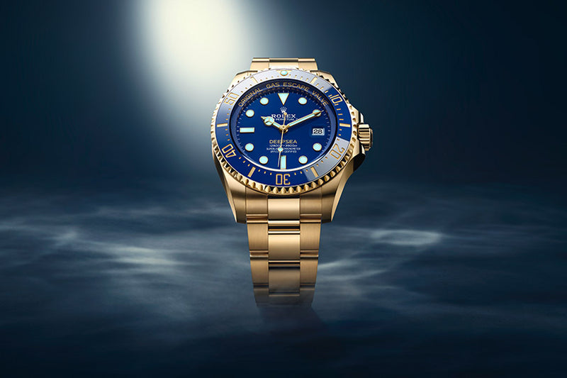 A gold Rolex watch with a blue dial is spotlighted against a dark, misty background.