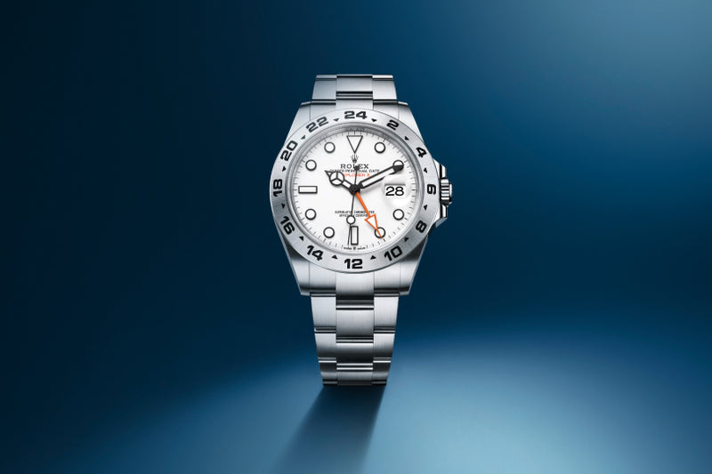 A silver Rolex Explorer II watch with a white dial and stainless steel bracelet on a blue gradient background.