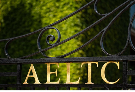 Black iron gate with gold letters AELTC, set against a blurred green leafy background.