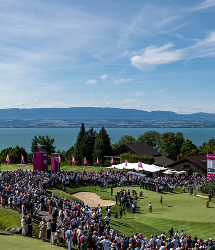 Large crowd watches a golf tournament by a lake, with mountains and blue sky in the background.