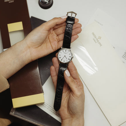 Two hands hold a watch with a black leather strap, behind the hands are an assortment of Patek Philippe branded papers and boxes