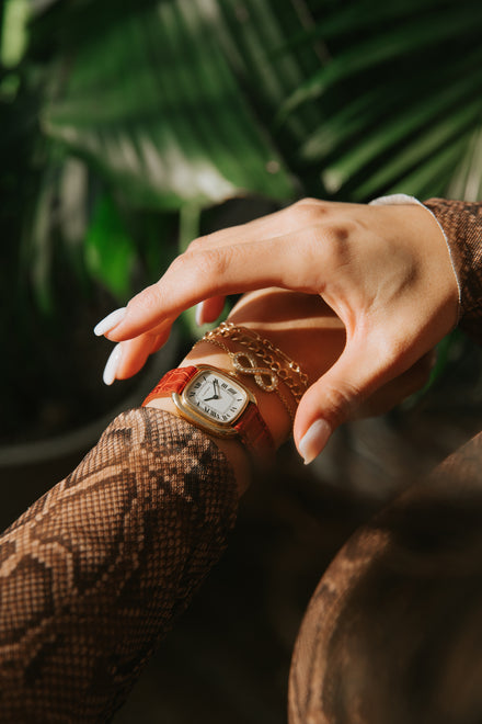 A hand with manicured nails wears a watch with a red band and gold bracelets, surrounded by green leaves.