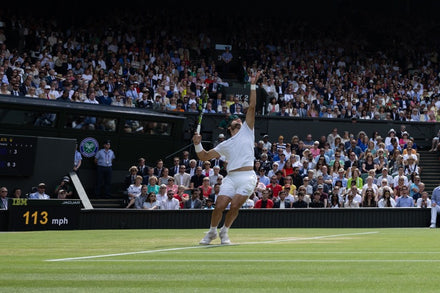 A tennis player serves on a grass court at Wimbledon, with a large crowd watching from the stands.