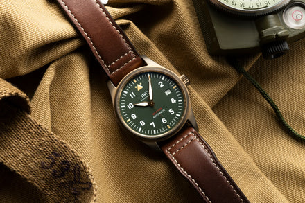 A wristwatch with a brown leather strap rests on tan fabric near a camera or instrument.
