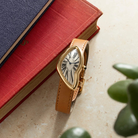 A gold, distorted watch with a tan leather strap rests on stacked books beside a green plant.