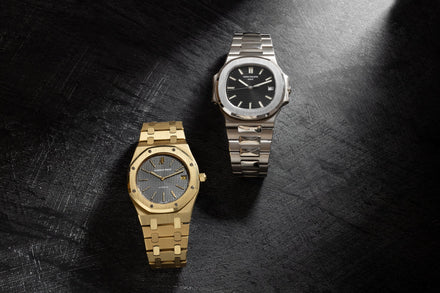 Two luxury watches, one gold and one silver, displayed on a textured black surface under a beam of light.