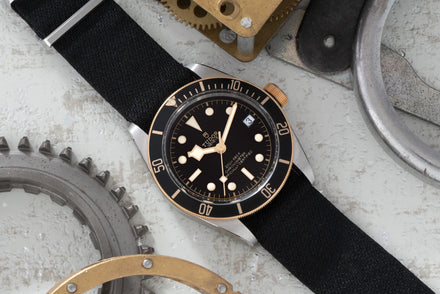 A black and gold wristwatch with a fabric strap, surrounded by metal gears on a textured surface.