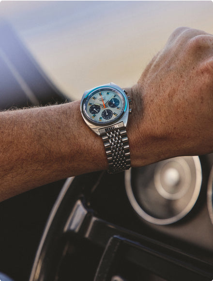 A close-up of a wristwatch with a blue face on a persons wrist, hand gripping a car steering wheel.