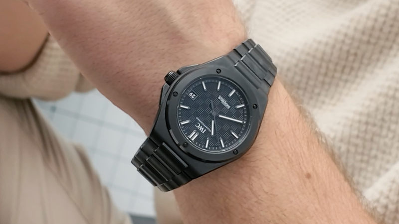 A black wristwatch with a textured face and metal band on a persons wrist, showing the time as 9:05.