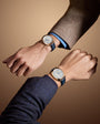 Two wrists wearing round watches with white dials, blue leather straps, and rose gold cases against a neutral tan background.
